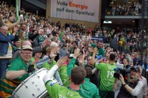 Eine jubelnde Menge in einer Sportarena, viele in grünen Trikots, jubelt, winkt und feiert mit den Spielern. Ein Schlagzeuger spielt im Vordergrund und ein Kameramann filmt die lebhafte Szene. Im Hintergrund hängt ein großes Transparent.