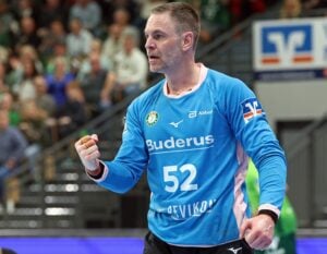 Ein männlicher Handballspieler in einem blauen Trikot mit der Nummer 52 ballt während eines Spiels jubelnd die Faust, während die Zuschauer im Hintergrund unscharf zu sehen sind.