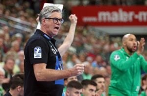 Ein Trainer mit Brille und schwarzem Polohemd gestikuliert energisch während eines Handballspiels, während im Hintergrund Spieler und eine jubelnde Menge zu sehen sind.