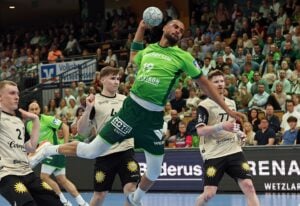 Ein Handballspieler in einem grünen Trikot springt in der Luft, um den Ball zu werfen. Er ist umgeben von gegnerischen Spielern in beigen Trikots, während im Hintergrund eine große Menschenmenge aufmerksam zusieht.
