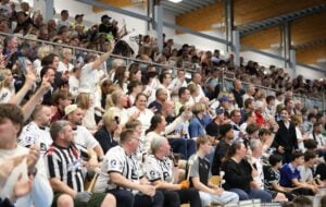 Eine große Menge begeisterter Zuschauer, viele in schwarzen und weißen Trikots und Hemden, sitzt auf den Tribünen in einer Sportarena und jubelt während eines Spiels und hebt die Arme.