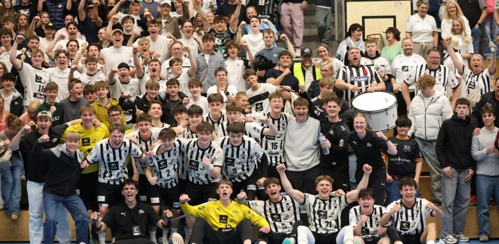 Eine große Gruppe aufgeregter junger Sportler in schwarz-weißen Trikots posiert zusammen mit jubelnden Fans in einer überfüllten Sporthalle. Einige Leute heben ihre Arme und lächeln begeistert.