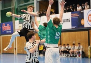 Ein Handballspieler in einem schwarz-weißen Trikot springt in die Luft, um den Ball zu schießen, während zwei Gegner in grünen Trikots versuchen, ihn zu blockieren. Im Hintergrund sind weitere Spieler und Zuschauer zu sehen.