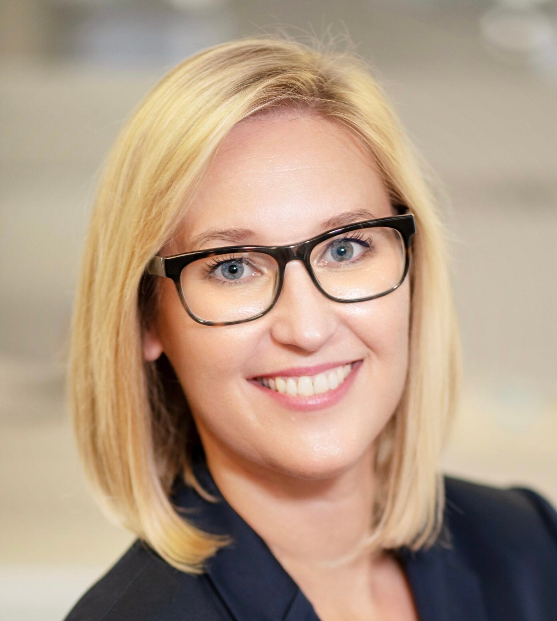 Eine Frau mit glattem blondem Haar und schwarzer Brille lächelt in die Kamera. Sie trägt einen marineblauen Blazer und steht in einer hell erleuchteten Halle. Sie stellt eines der Mitglieder der THW Kiel Geschäftsführung dar.