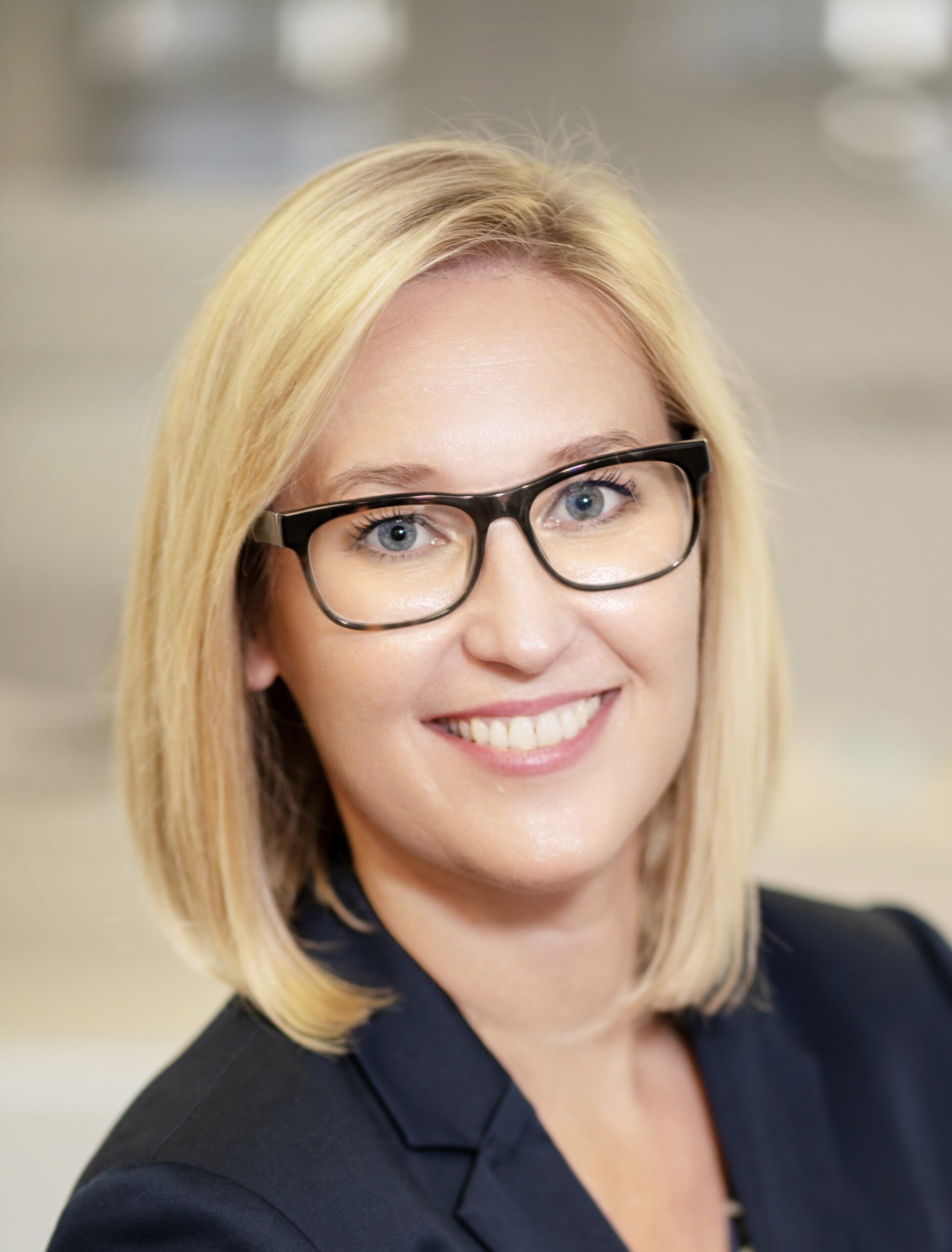 Anja Eine Frau mit glattem blondem Haar und schwarzer Brille lächelt in die Kamera. Sie trägt einen marineblauen Blazer und steht in einer hell erleuchteten Halle. Sie stellt eines der Mitglieder der THW Kiel Geschäftsführung dar.