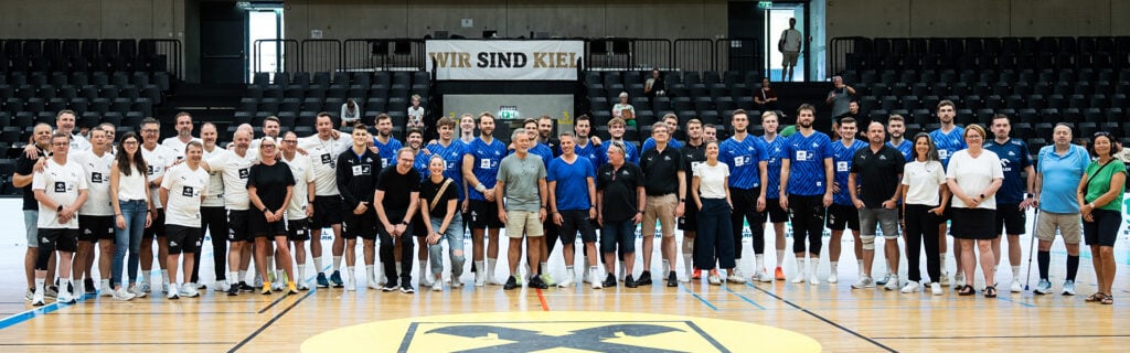 Eine große Gruppe von Menschen, darunter Spieler in Sportuniformen und andere in Freizeitkleidung, stehen gemeinsam auf einem Hallenplatz. Hinter ihnen sind leere Tribünen und ein Transparent mit der Aufschrift WIR SIND KIEL zu sehen.