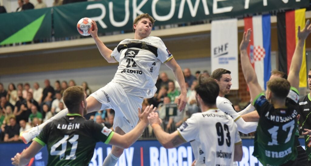 Ein Handballspieler in einem weißen Trikot springt in die Luft, um den Ball zu werfen, umgeben von Gegnern in grünen Trikots, während eines intensiven Spiels, das die Zuschauer von der Tribüne aus verfolgen.