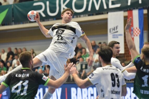 Ein Handballspieler in einem weißen Trikot springt in die Luft, um den Ball zu werfen, umgeben von Gegnern in grünen Trikots, während eines intensiven Spiels, das die Zuschauer von der Tribüne aus verfolgen.