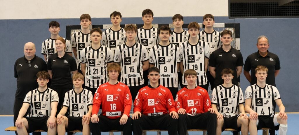 Ein Gruppenfoto einer Jugendhandballmannschaft in schwarz-weiß gestreiften Trikots mit vier Trainern in schwarzen Hemden. Die Spieler stehen und sitzen in Reihen auf einer Bank in einer Sporthalle.