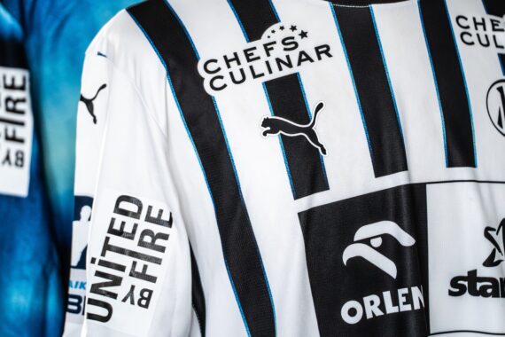 Nahaufnahme von Sporttrikots mit verschiedenen Sponsorenlogos, darunter Chefs Culinar, United by Fire, Orlen und Puma, in Schwarz und Weiß mit blauen Akzenten. Im Hintergrund ist ein Teil eines anderen blauen Trikots zu sehen.