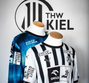 Zwei Schaufensterpuppen zeigen Handballtrikots des THW Kiel, eines in Blau mit schwarzen Akzenten, das andere in Weiß mit schwarzen Längsstreifen. An der Wand dahinter ist das Logo des THW Kiel zu sehen.