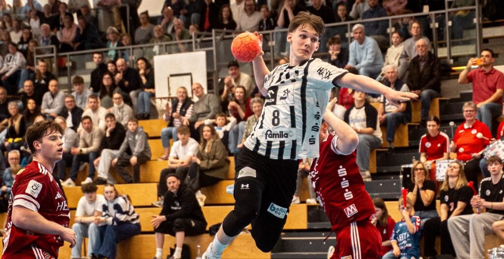 Ein Handballspieler in einem weiß-schwarzen Trikot springt in die Luft, um den Ball zu werfen, während zwei Spieler in roten Trikots den Ball abwehren. Die Zuschauer auf der Tribüne im Hintergrund verfolgen das Geschehen aufmerksam.