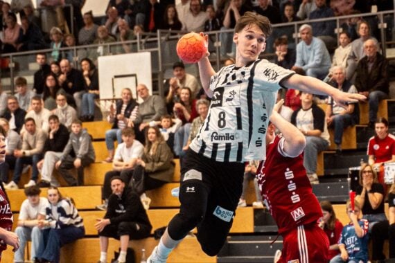 Ein Handballspieler in einem weiß-schwarzen Trikot springt in die Luft, um den Ball zu werfen, während zwei Spieler in roten Trikots den Ball abwehren. Die Zuschauer auf der Tribüne im Hintergrund verfolgen das Geschehen aufmerksam.