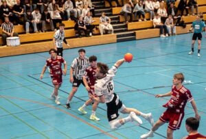 Ein Handballspieler in einem weiß-schwarzen Trikot springt, um den Ball in Richtung Tor zu werfen, während Verteidiger in roten Trikots versuchen, ihn abzuwehren. Die Zuschauer schauen von einer hölzernen Tribüne im Hintergrund zu.