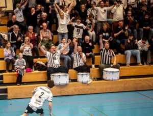 Fans in schwarz-weißen Hemden jubeln und trommeln auf überfüllten Tribünen bei einer Hallensportveranstaltung, während ein Spieler auf dem Spielfeld mit der Nummer 2 auf seinem Trikot zum Publikum schaut. Die Atmosphäre ist lebhaft und energiegeladen.