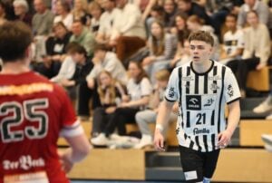 Ein junger männlicher Handballspieler in einem schwarz-weiß gestreiften Trikot mit der Nummer 21 blickt konzentriert auf das Spielfeld, während ein anderer Spieler in einem roten Trikot mit der Nummer 25 im Vordergrund steht. Die Zuschauer schauen von der Tribüne aus zu.