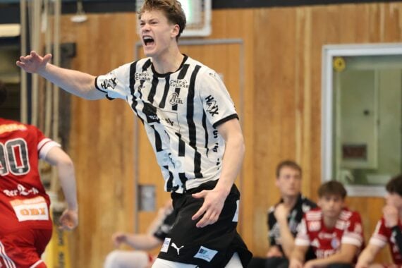 Ein junger männlicher Handballspieler in einem weiß-schwarzen Trikot schreit energisch während eines Spiels. Gegner in roten Trikots und Mannschaftskameraden sitzen auf einer Bank im Hintergrund in einer Sporthalle.