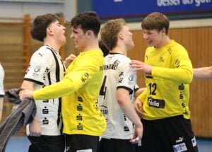 Vier junge männliche Sportler in gelben und weißen Trikots feiern gemeinsam, lächelnd und lachend auf einem Hallenplatz. Zwei tragen gelb, zwei weiß; sie wirken nach einem Spiel fröhlich, stehen dicht beieinander und unterhalten sich enthusiastisch.