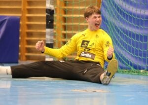Ein Handballtorwart in einem gelben Trikot sitzt mit gespreizten Beinen und geballten Fäusten auf dem Boden und feiert ausgelassen vor einem Tornetz in einer Sporthalle.