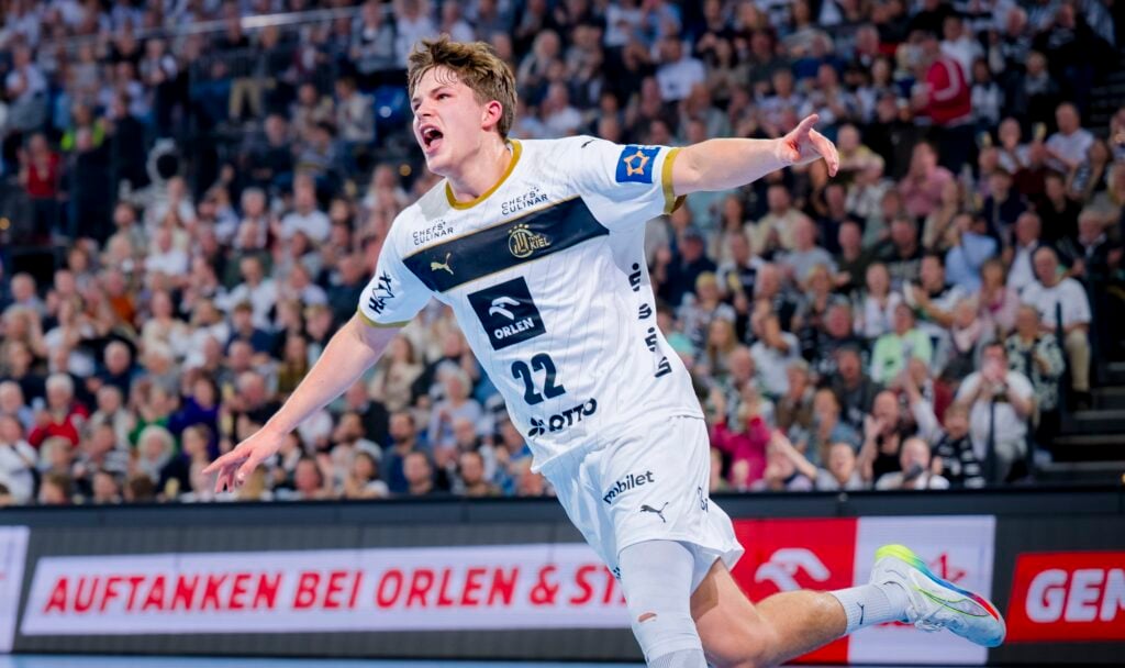 Ein männlicher Handballspieler in einem weißen Trikot mit der Nummer 22 jubelt mit ausgestreckten Armen auf einem Hallenplatz. Im Hintergrund ist eine unscharfe Zuschauermenge zu sehen.