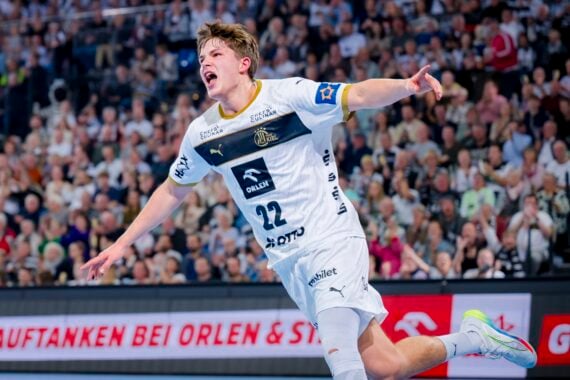 Ein männlicher Handballspieler in einem weißen Trikot mit der Nummer 22 jubelt mit ausgestreckten Armen auf einem Hallenplatz. Im Hintergrund ist eine unscharfe Zuschauermenge zu sehen.