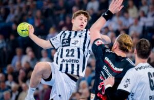 Ein Handballspieler in einem weiß-schwarz gestreiften Trikot springt, um einen gelben Ball zu werfen, während ein Verteidiger in einem schwarzen Trikot den Arm hebt, um ihn abzublocken, während im Hintergrund eine verschwommene Menschenmenge zu sehen ist.