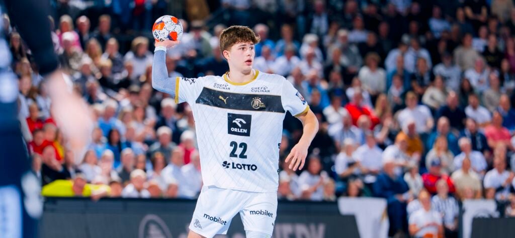 Ein männlicher Handballspieler in einem weißen Trikot mit der Nummer 22 hält einen Ball und bereitet sich auf den Wurf vor, während er auf einem Spielfeld mit einer verschwommenen Menschenmenge im Hintergrund steht.