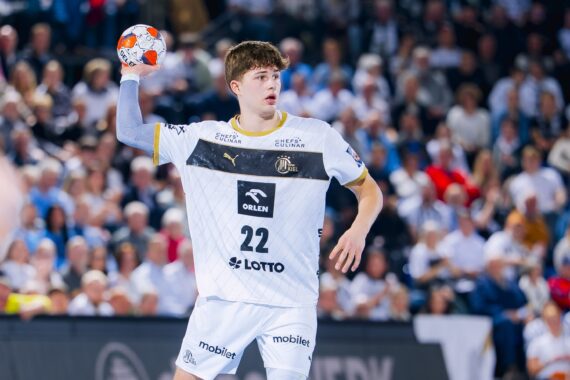 Ein männlicher Handballspieler in einem weißen Trikot mit der Nummer 22 hält einen Ball und bereitet sich auf den Wurf vor, während er auf einem Spielfeld mit einer verschwommenen Menschenmenge im Hintergrund steht.