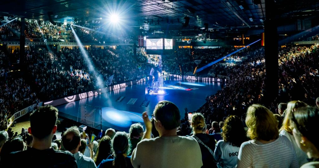 Eine große Menschenmenge in einer Halle sieht zu, wie ein Scheinwerfer die Spieler auf dem Handballfeld anstrahlt. Das Stadion ist hell erleuchtet und voller Aufregung.