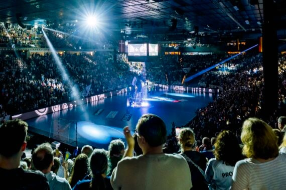 Eine große Menschenmenge in einer Halle sieht zu, wie ein Scheinwerfer die Spieler auf dem Handballfeld anstrahlt. Das Stadion ist hell erleuchtet und voller Aufregung.