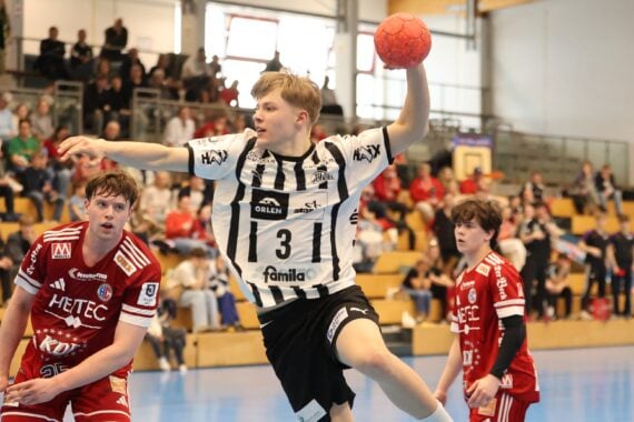 Ein Handballspieler in einem schwarz-weiß gestreiften Trikot springt in der Luft, um den Ball zu werfen. Zwei Spieler in roten Trikots stehen in der Nähe, die Zuschauer schauen von der Tribüne in einer hell erleuchteten Turnhalle zu.