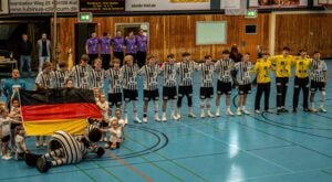 Eine Handballmannschaft in schwarz-weißen Trikots steht mit verschränkten Armen in einer Reihe auf einem Hallenplatz. Zwei Mannschaftsmaskottchen und mehrere Kinder halten eine große deutsche Flagge, während Trainer und Betreuer im Hintergrund stehen.