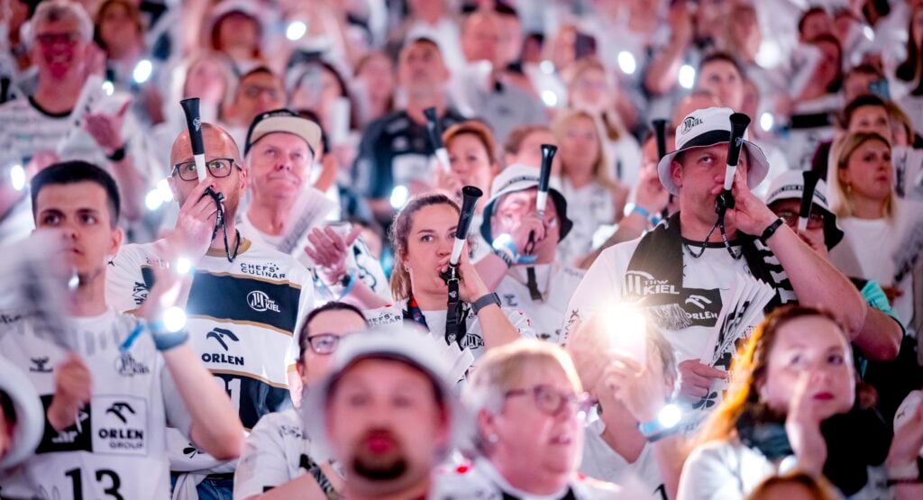 Eine große Menge begeisterter, meist weiß gekleideter Sportfans hält leuchtende Smartphones in die Höhe und bläst in Vuvuzelas, um eine lebhafte und energiegeladene Atmosphäre bei einer Hallenveranstaltung zu schaffen.