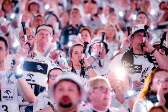 Eine große Menge begeisterter, meist weiß gekleideter Sportfans hält leuchtende Smartphones in die Höhe und bläst in Vuvuzelas, um eine lebhafte und energiegeladene Atmosphäre bei einer Hallenveranstaltung zu schaffen.