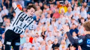 Ein Handballspieler in einem schwarz-weißen Trikot bereitet sich darauf vor, den Ball in Richtung Tor zu werfen, während ein Torwart sich bereit macht, ihn abzublocken. Im Hintergrund ist eine unscharfe Zuschauermenge zu sehen, die aufmerksam zusieht.