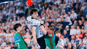 Ein Handballspieler in einem weißen Trikot springt, um den Ball zu schießen, während zwei Verteidiger in grünen Trikots versuchen, ihn während eines intensiven Spiels zu blockieren, während im Hintergrund eine unscharfe Menge zusieht.