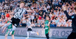 Ein Handballspieler in einem schwarz-weiß gestreiften Trikot springt mit einem roten Ball in der Hand auf ein Tor zu, während sich ein Torwart darauf vorbereitet, den Ball zu blocken. Die Szene spielt sich in einer überfüllten Halle ab, in der Zuschauer zuschauen.