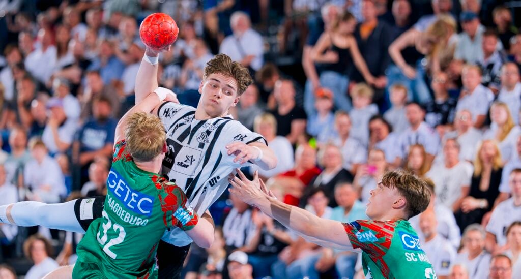 Ein Handballspieler in einem schwarz-weißen Trikot springt, um den Ball zu werfen, während zwei Verteidiger in grünen Trikots versuchen, ihn zu blockieren. Die Aktion findet vor einem überfüllten Zuschauerraum statt.