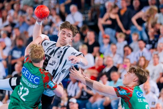 Ein Handballspieler in einem schwarz-weißen Trikot springt, um den Ball zu werfen, während zwei Verteidiger in grünen Trikots versuchen, ihn zu blockieren. Die Aktion findet vor einem überfüllten Zuschauerraum statt.