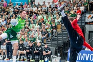 Ein Handballspieler im grünen Trikot springt zum Schuss, während ein Torhüter in Rot sein Bein hoch streckt, um das Spiel zu verhindern, während die Zuschauer von der Tribüne aus aufmerksam zusehen.