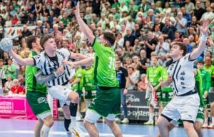 Ein spannender Moment in einem Handballspiel zeigt die Spieler zweier Mannschaften, eine in Grün und eine in Weiß, die um den Ball kämpfen, während die Zuschauer im Hintergrund aufmerksam zusehen.