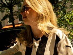 Eine Frau mit langen blonden Haaren und Sonnenbrille trägt ein schwarz-weiß gestreiftes Sporttrikot. Sie steht im Freien im Sonnenlicht mit Bäumen und einem Auto im Hintergrund.