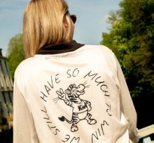 Eine Person mit langen blonden Haaren steht im Freien und wendet sich ab. Sie trägt eine weiße Jacke mit einem Cartoon-Tiger, der ein Friedenszeichen gibt, und dem Text "Wir haben noch so viel zu gewinnen", der in einem kreisförmigen Muster aufgedruckt ist. Es sind Bäume und ein Gebäude zu sehen.
