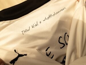 Nahaufnahme eines weißen Sporttrikots mit schwarzem Kragen, das den handgeschriebenen Text THW Kiel x whattabeanwears in der Nähe des Halsausschnitts und einen Teil des Puma-Logos auf der linken Seite zeigt.