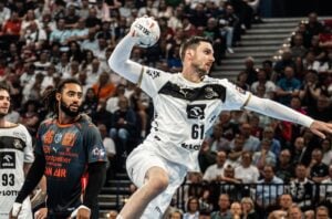 Ein Handballspieler in einem weiß-schwarzen Trikot springt und bereitet sich auf den Wurf vor, während ein gegnerischer Spieler in einem dunkelgrauen und orangefarbenen Trikot zuschaut. Das Spiel findet in einer überfüllten Halle statt.