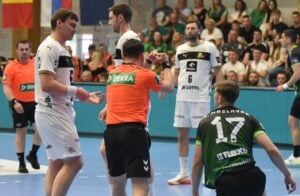 Ein Schiedsrichter in einem orangefarbenen Hemd zeigt einem Spieler in einem weißen Trikot während eines Hallenhandballspiels eine Handbewegung, während andere Spieler und Zuschauer das Geschehen beobachten.