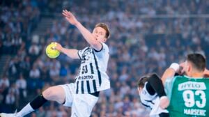 Ein männlicher Handballspieler in einem schwarz-weißen Trikot befindet sich in der Luft, um einen gelben Ball zu werfen, während seine Gegner in grünen Trikots ihn verteidigen. Der Hintergrund zeigt eine unscharfe Menschenmenge in einer vollen Arena.