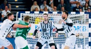 Vier männliche Handballspieler, drei in weiß-schwarz gestreiften Trikots und einer in grün, spielen aktiv auf einem Hallenplatz, während im Hintergrund eine Menschenmenge zusieht.