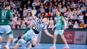 Ein Handballspieler in einem schwarz-weiß gestreiften Trikot springt, um den Ball zu schießen, während er von gegnerischen Spielern in grünen Trikots umringt ist, während die Zuschauer in einer Halle zuschauen.