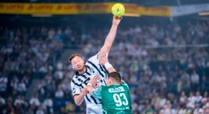 Ein Handballspieler in einem schwarz-weiß gestreiften Trikot springt und holt aus, um einen gelben Ball zu werfen, während ein Verteidiger in einem grünen Trikot mit der Nummer 93 versucht, ihn abzuwehren. Die Zuschauer im Hintergrund schauen gespannt zu.